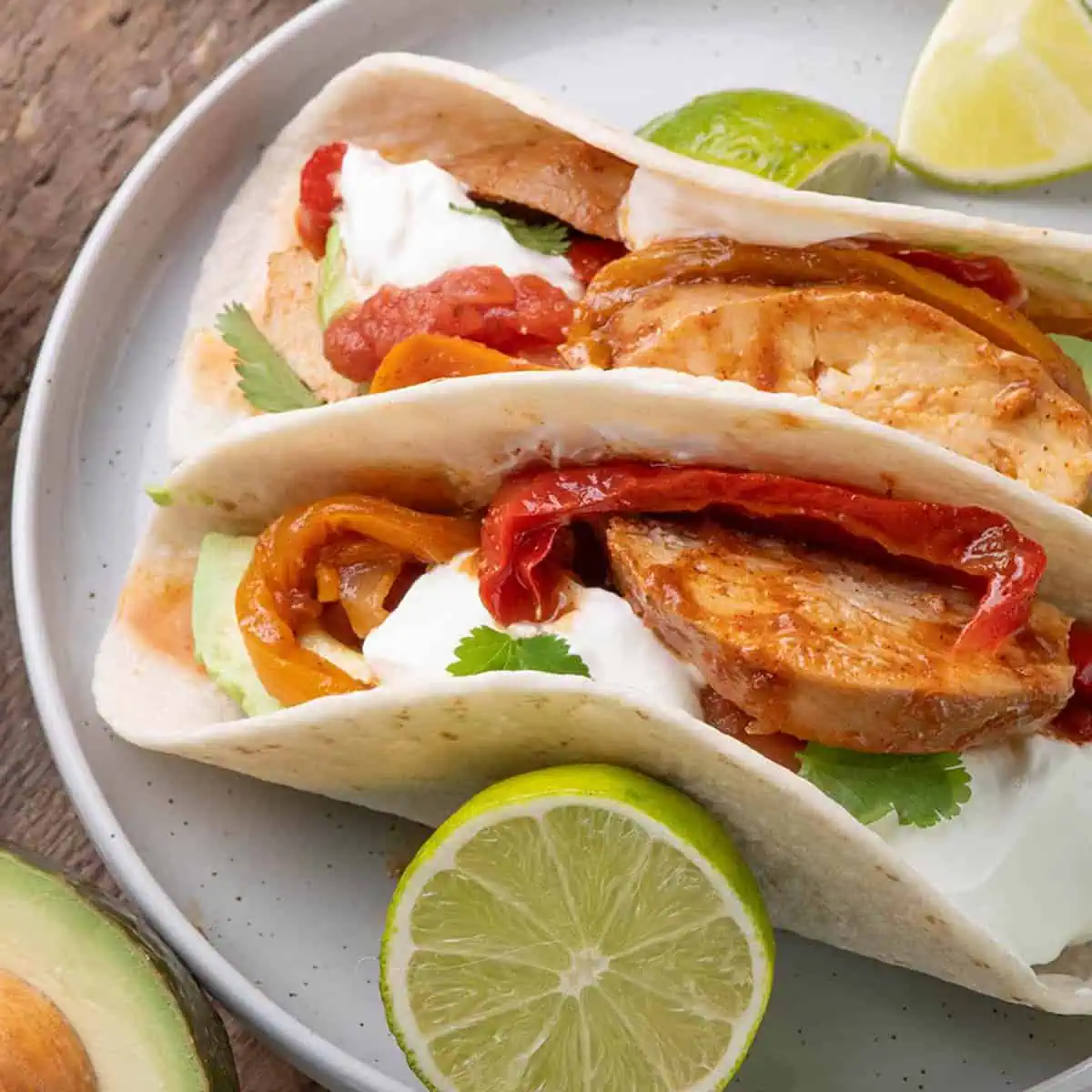 Chicken fajitas in soft flour tortillas with sautéed peppers, sour cream, and fresh cilantro, served on a gray plate with lime wedges.
