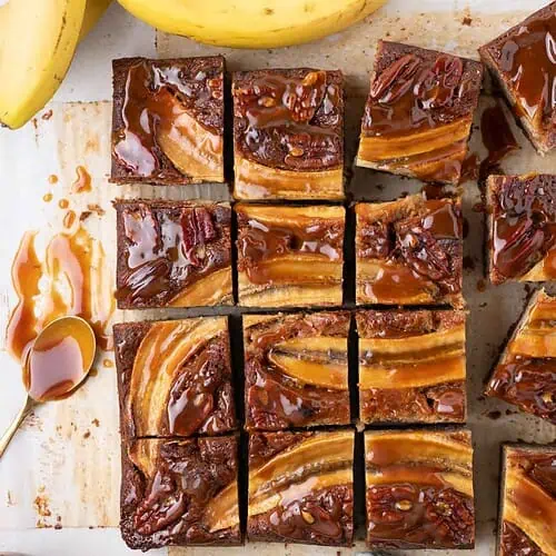 Overhead view of sliced banana bread bars topped with bananas, pecans, and glossy caramel, with a rustic knife, spoon, and fresh bananas in the frame