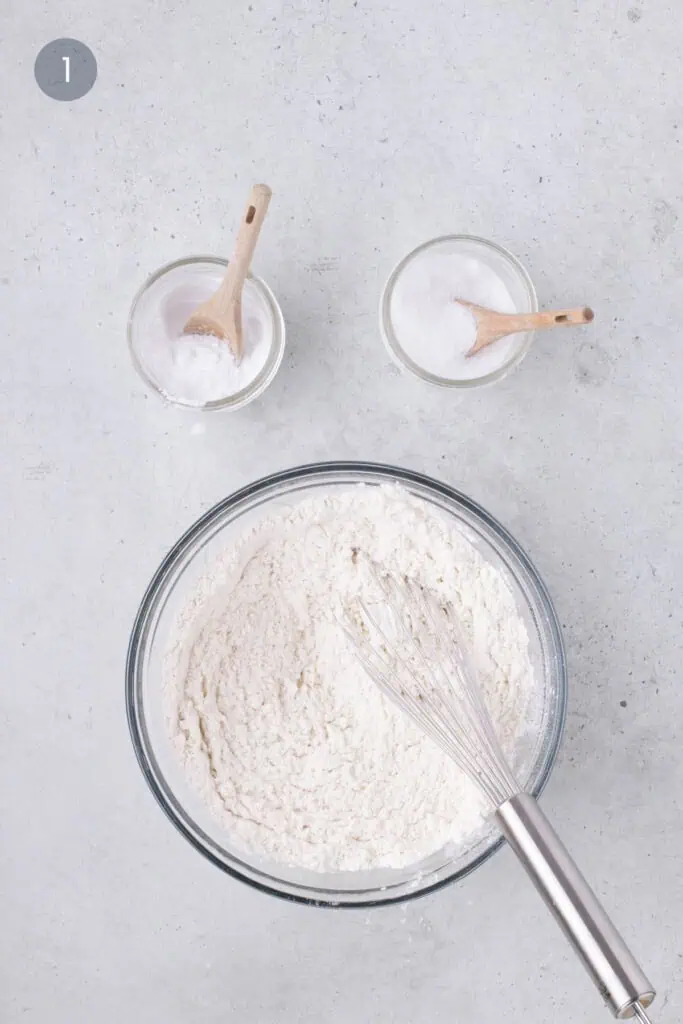 Flour, salt, and baking soda whisked together in a glass bowl on a light countertop.