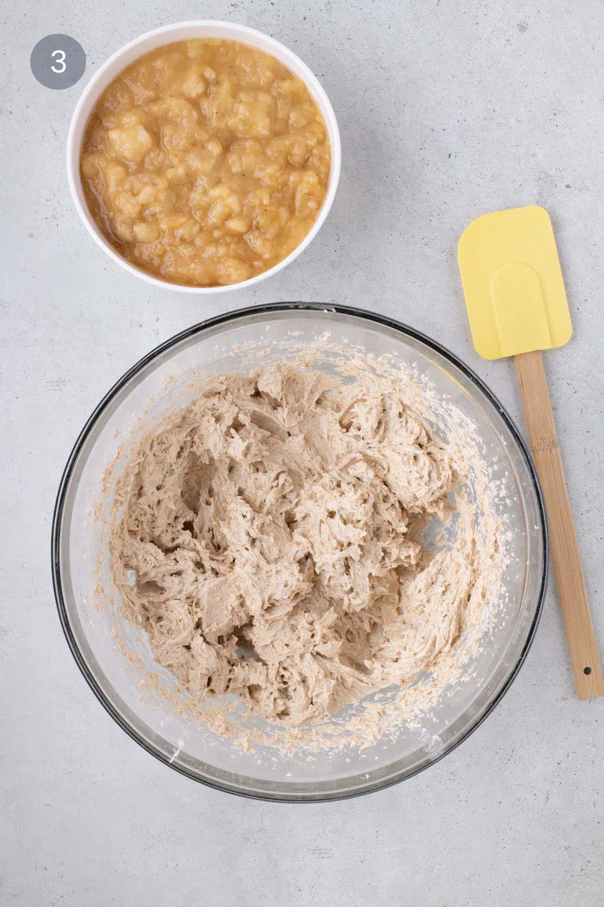 Mixing bowl of batter next to mashed bananas and a yellow spatula.