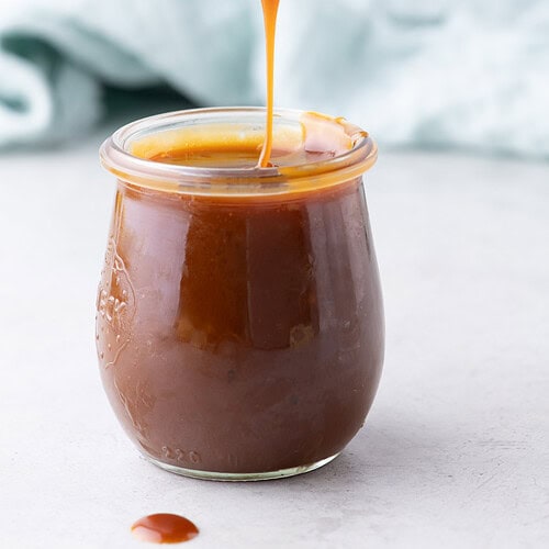 Golden spoon lifting salted caramel sauce over a glass jar with caramel dripping back into the jar.