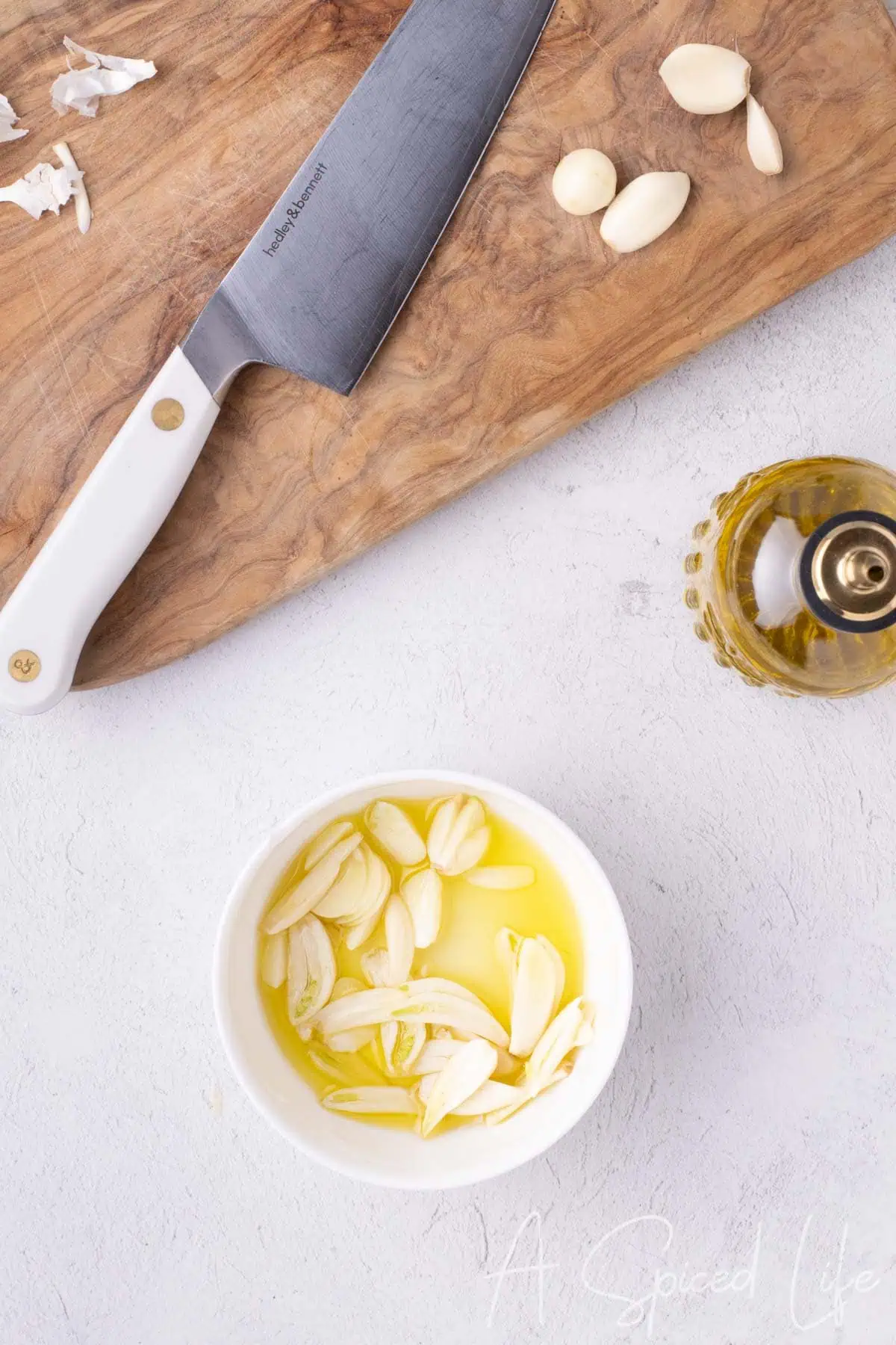 Sliced garlic soaking in olive oil next to a chef’s knife on a cutting board