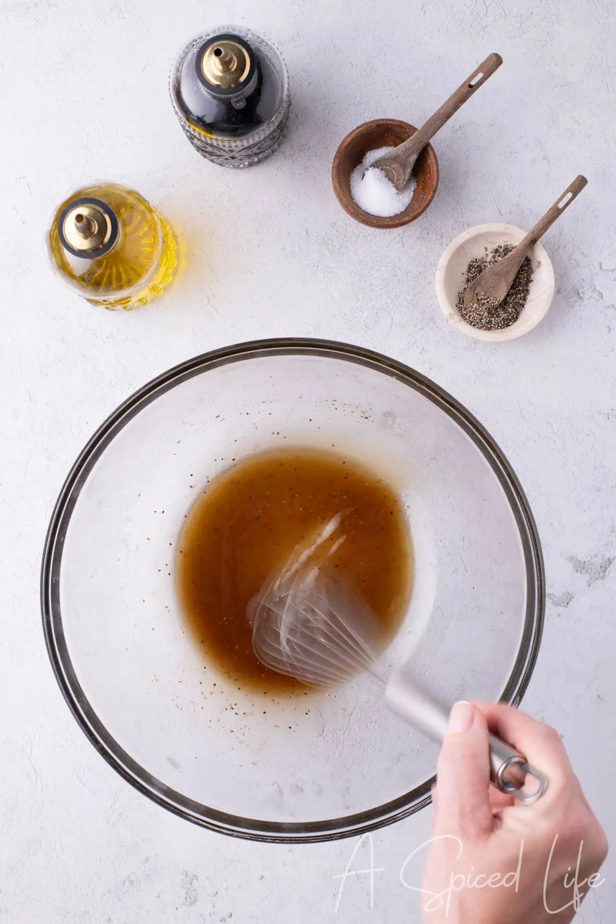 Hand whisking balsamic vinegar and olive oil in a glass bowl with salt and pepper nearby.