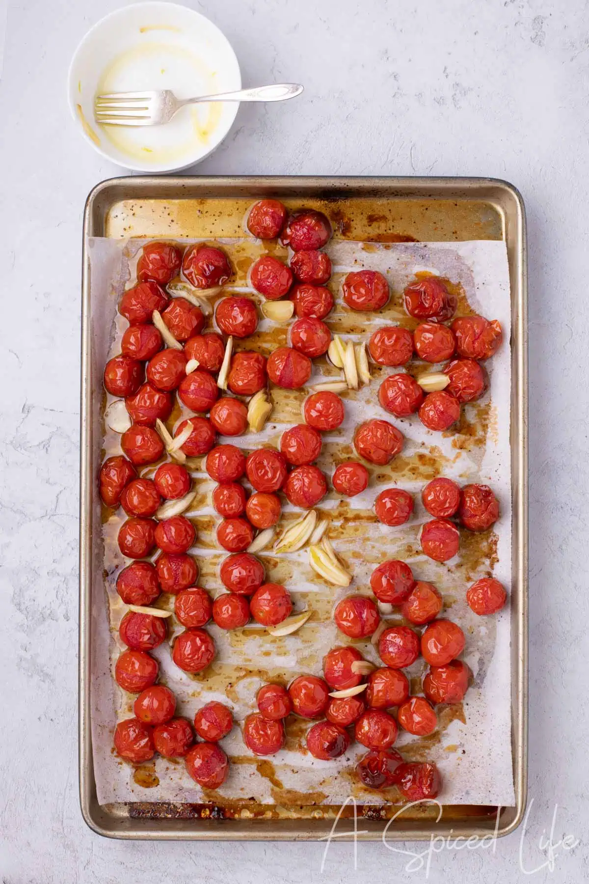Partially roasted cherry tomatoes with sliced garlic added on a parchment-lined sheet pan.