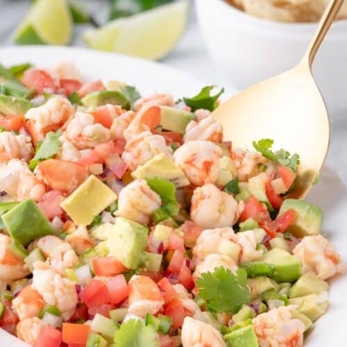 Shrimp ceviche on a white serving plate with a gold spoon, surrounded by lime wedges and tortilla chips.