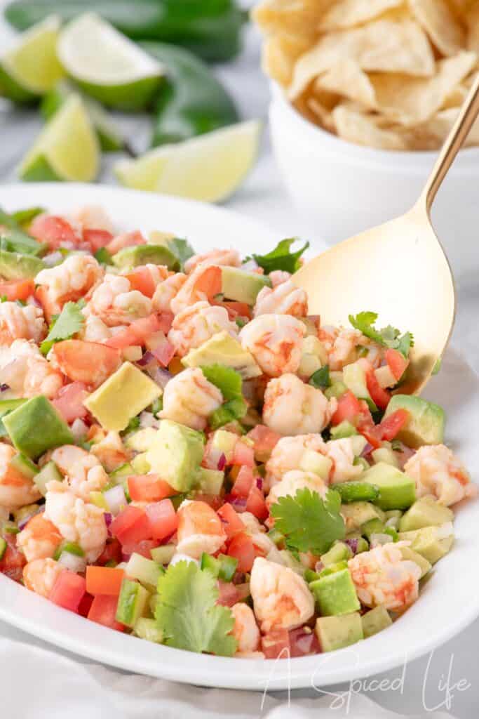 Shrimp ceviche on a white serving plate with a gold spoon, surrounded by lime wedges and tortilla chips.