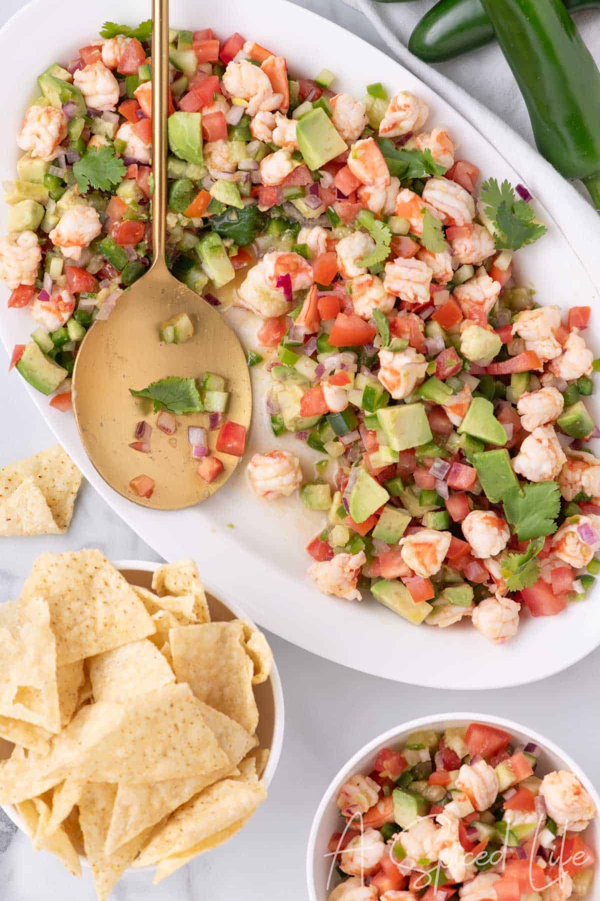 Bright shrimp ceviche with avocado and tomatoes served on a platter with a gold spoon and chips nearby.