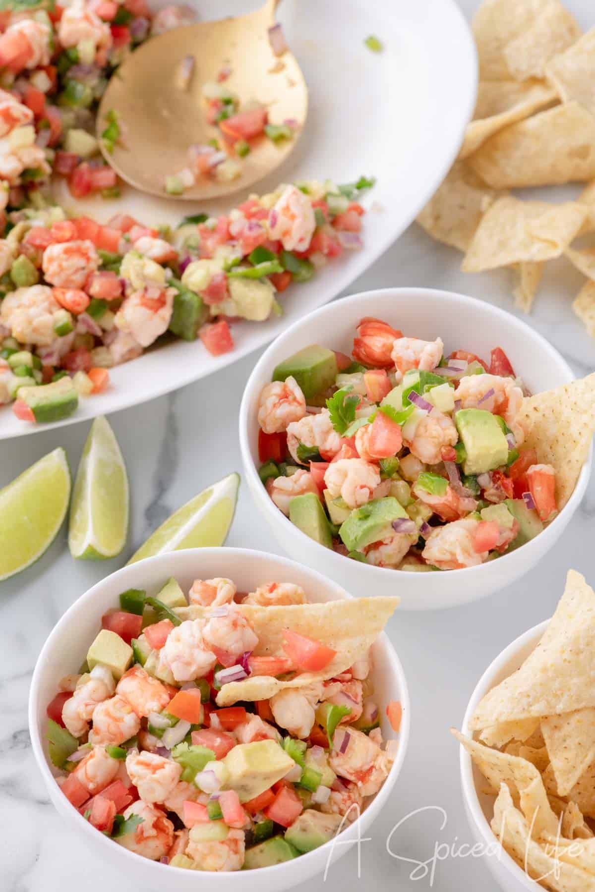 Overhead view of two small bowls of shrimp ceviche with lime wedges, chips, and a serving plate nearby.