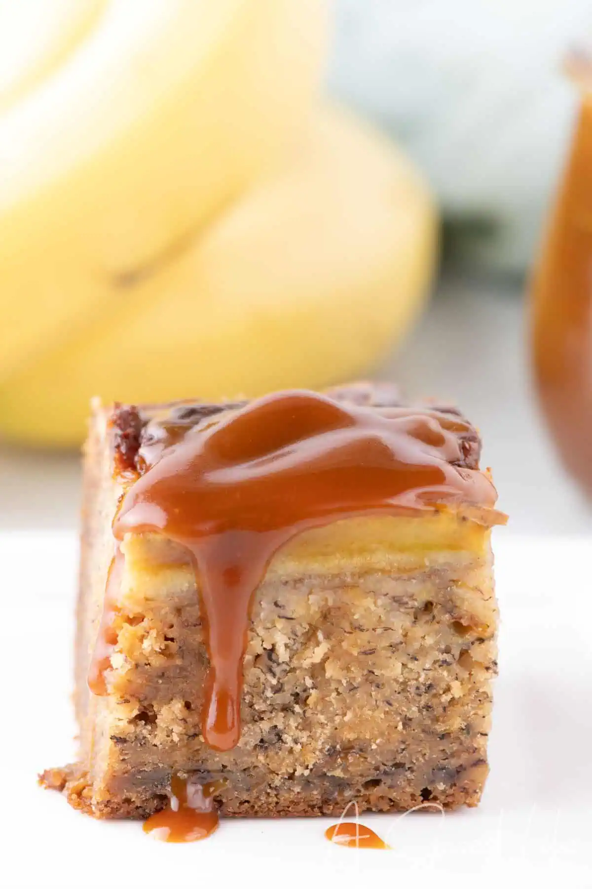 Close-up of a gooey banana bread bar topped with a thick layer of golden salted caramel, with bananas blurred in the background.