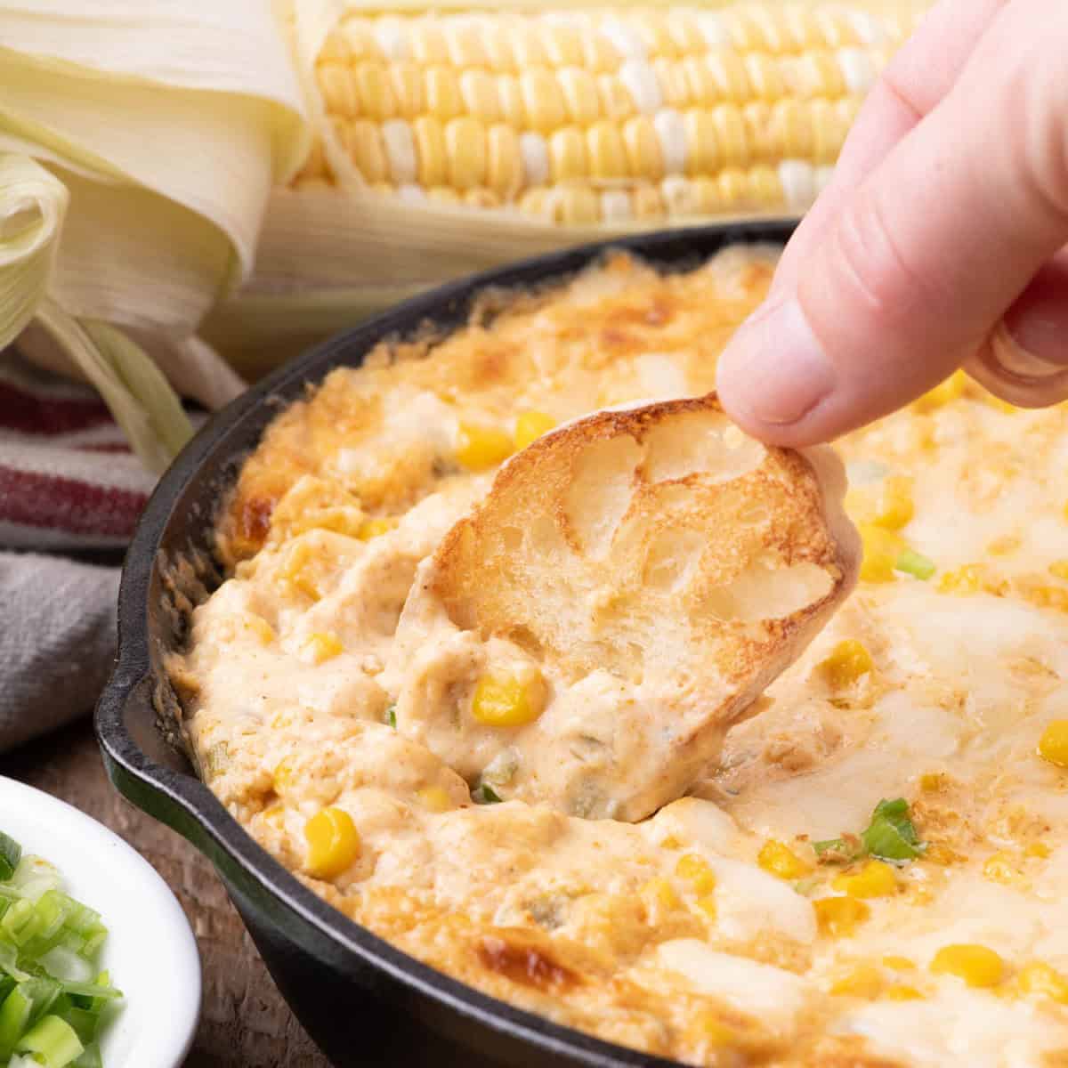 Hand dipping toasted baguette into hot corn and cream cheese dip in a cast-iron skillet, with fresh corn cobs in the background.