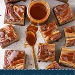 Overhead photo of banana bread bars topped with salted caramel sauce and pecans, arranged around a jar of caramel with a spoon.