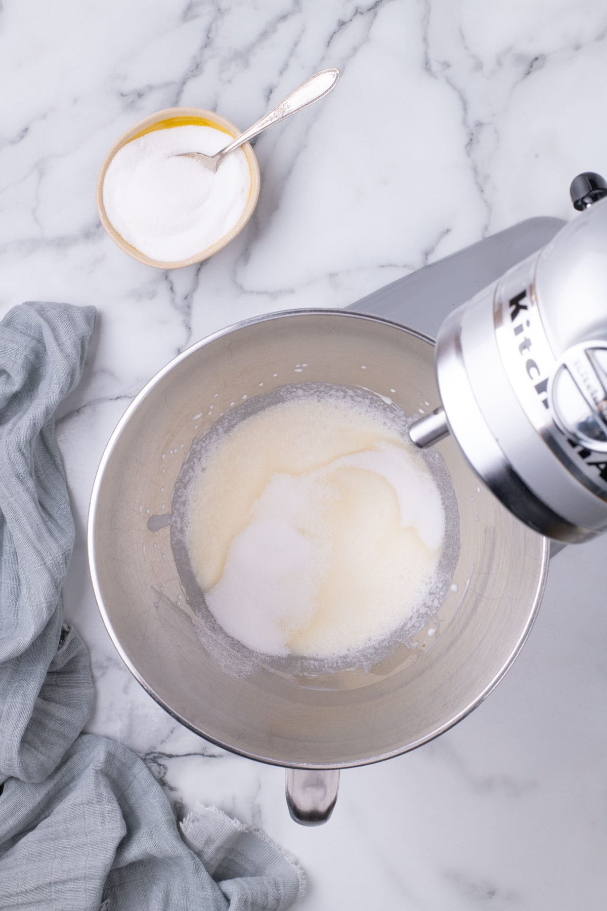 Sugar added to whipped cream mixture in a stand mixer bowl.