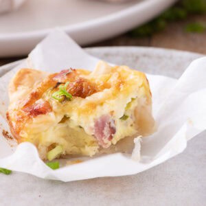 Close-up of a mini quiche with puff pastry, cottage cheese, ham, and green onions on a plate with parchment paper