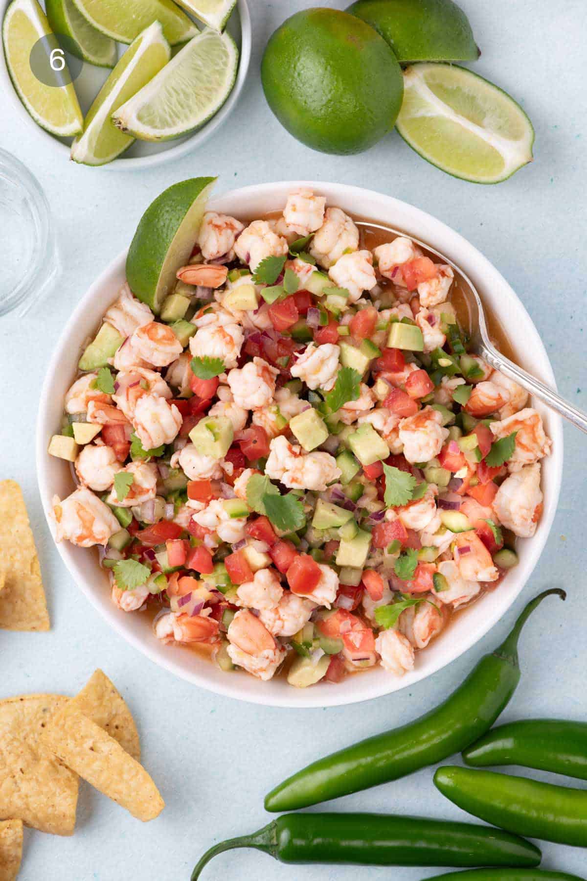 Bowl filled with shrimp, avocado, tomato, red onion, and cilantro marinating in lime juice, surrounded by fresh limes, serrano peppers, and tortilla chips.