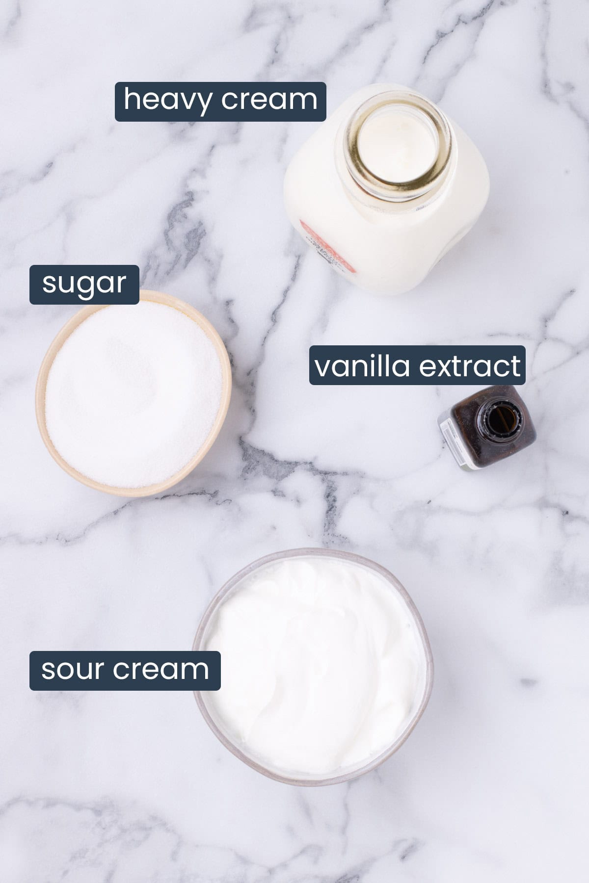 Overhead view of ingredients for Chantilly cream including heavy cream, sugar, vanilla, and sour cream on a marble surface.