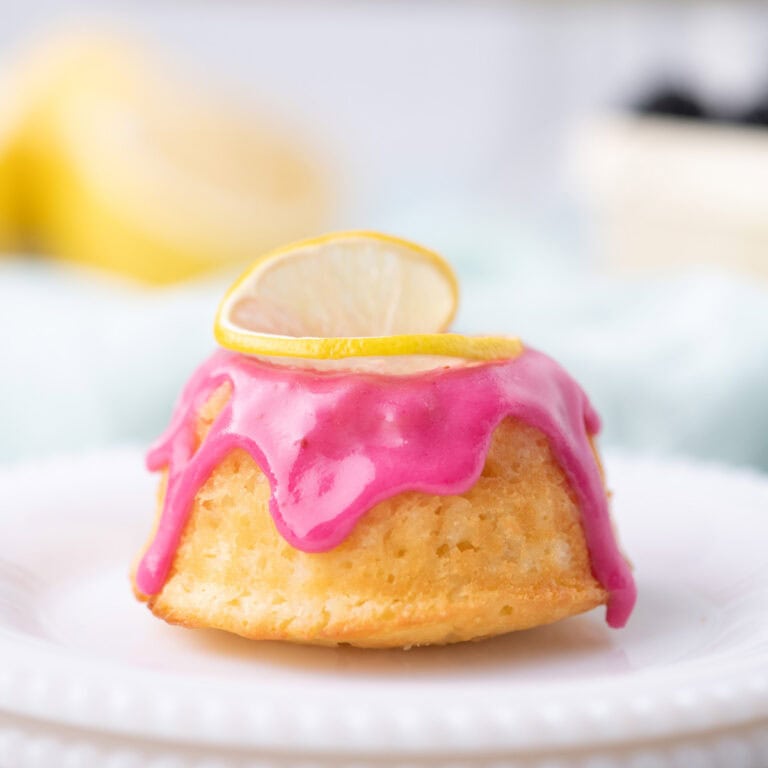 Lemon mini bundt cake topped with bright pink blackberry glaze and a lemon slice garnish on a white plate.