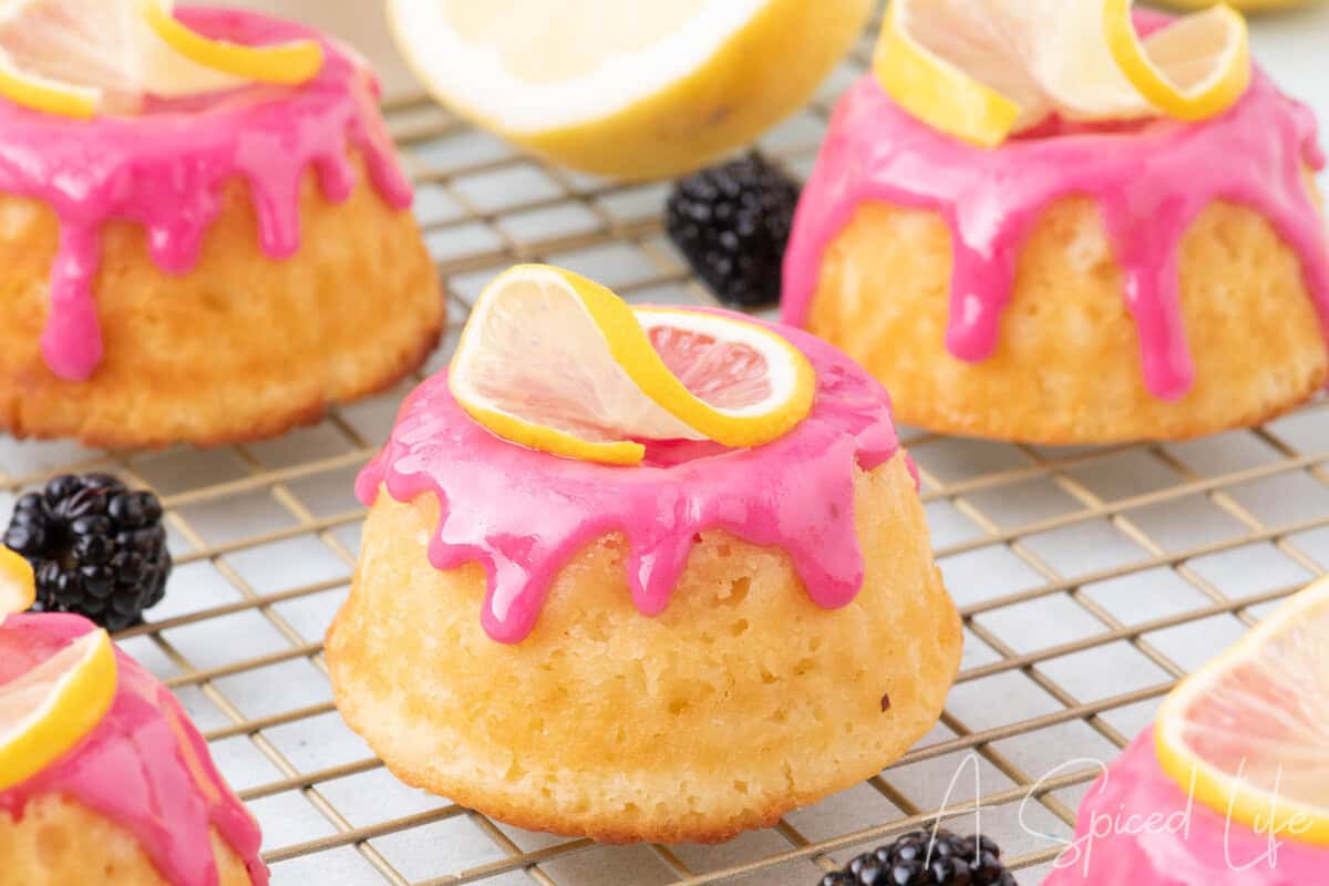 Close-up of lemon mini bundt cakes topped with bright pink blackberry glaze and lemon slices on a cooling rack.