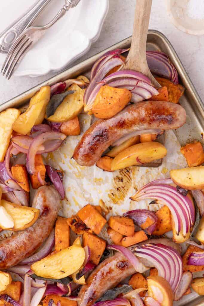 Roasted sausages with sweet potatoes, apples, and onions on a rustic baking sheet.