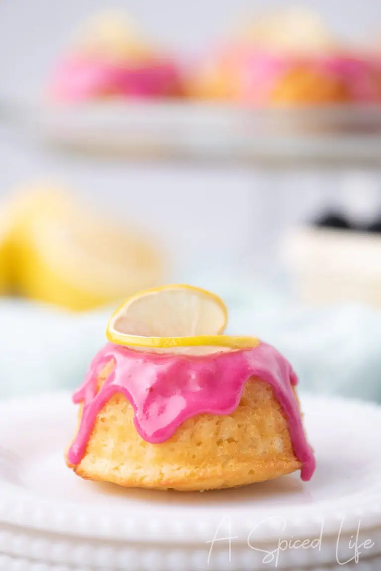 Lemon mini bundt cake topped with bright pink blackberry glaze and a lemon slice, served on stacked white plates with blurred background.