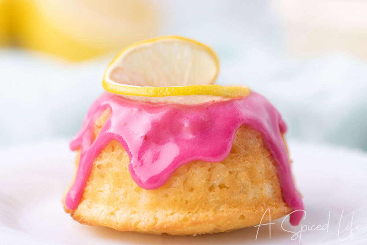 Close-up of a lemon mini bundt cake with vibrant pink blackberry glaze dripping down the sides, topped with a thin lemon slice.