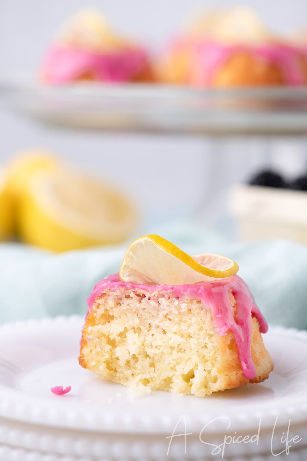Slice of lemon mini bundt cake with bright pink blackberry glaze and a lemon slice garnish, showing moist crumb texture on a white plate.