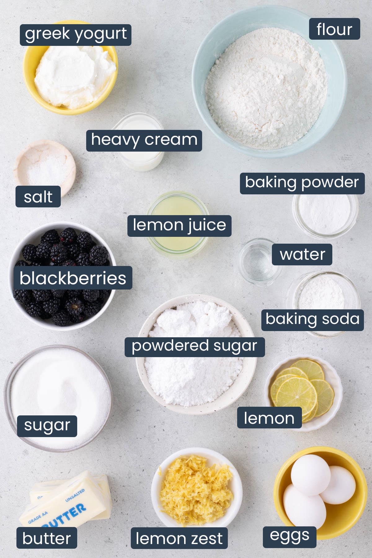 Overhead view of flour, sugar, butter, eggs, blackberries, lemon, yogurt, and other baking ingredients arranged on a light surface