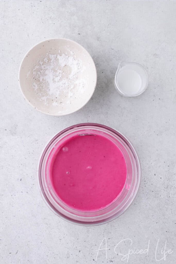 Bright pink blackberry glaze in a bowl with powdered sugar and milk nearby