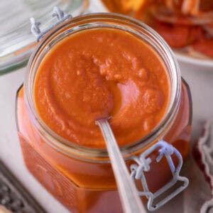 A glass jar of homemade tomato sauce with butter and San Marzano tomatoes, with a spoon and spaghetti in the background.