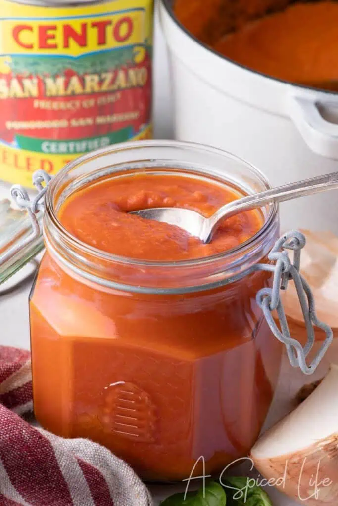 A glass jar filled with homemade San Marzano tomato sauce, with a spoon and a can of certified tomatoes in the background.