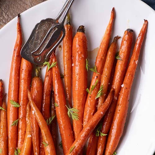 Maple Glazed Carrots served on a white platter with fresh carrot fronds and a buttery maple syrup glaze.