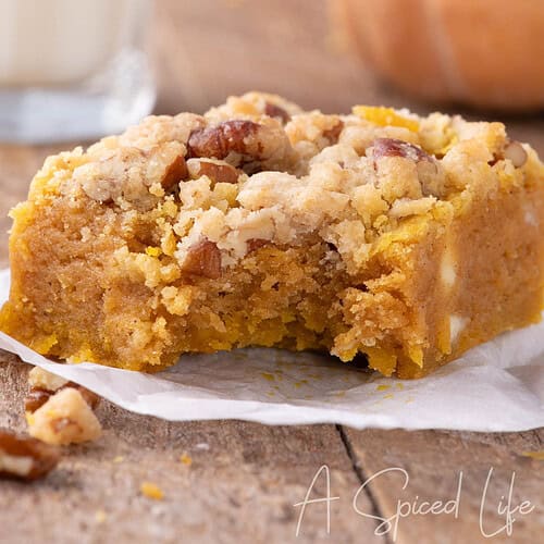 Close-up of a pumpkin blondie with a bite taken out, showing soft texture and pecan crumble topping, with pumpkins and milk in the background.