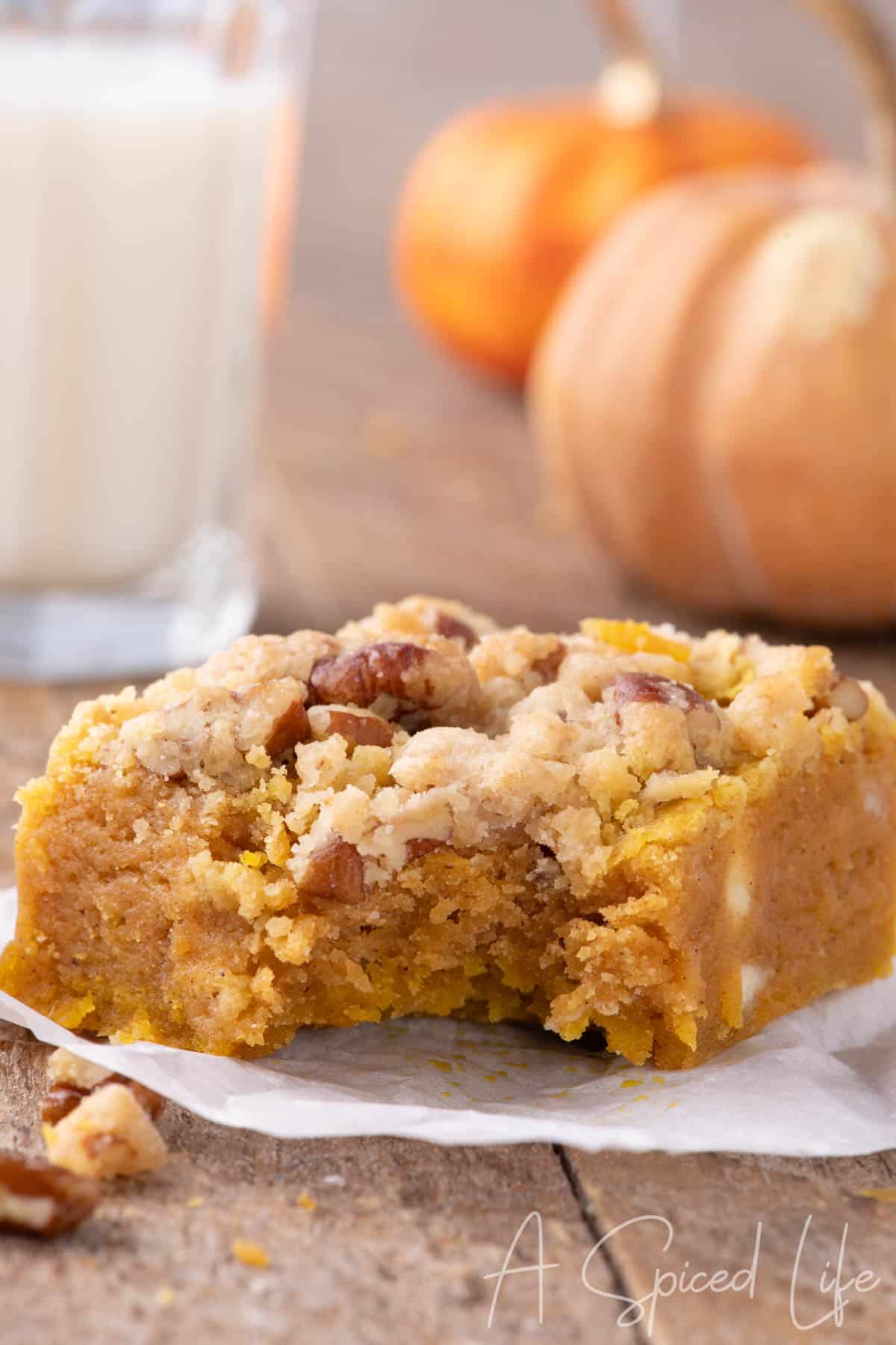 Close-up of a pumpkin blondie with a bite taken out, showing soft texture and pecan crumble topping, with pumpkins and milk in the background.