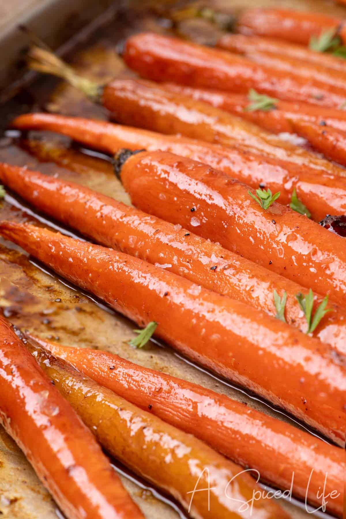 Close-up of Maple Glazed Carrots roasted until glossy with sea salt and fresh carrot fronds on a sheet pan.