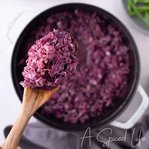Wooden spoon lifting German braised red cabbage from a Dutch oven, with roast chicken and herbs in background