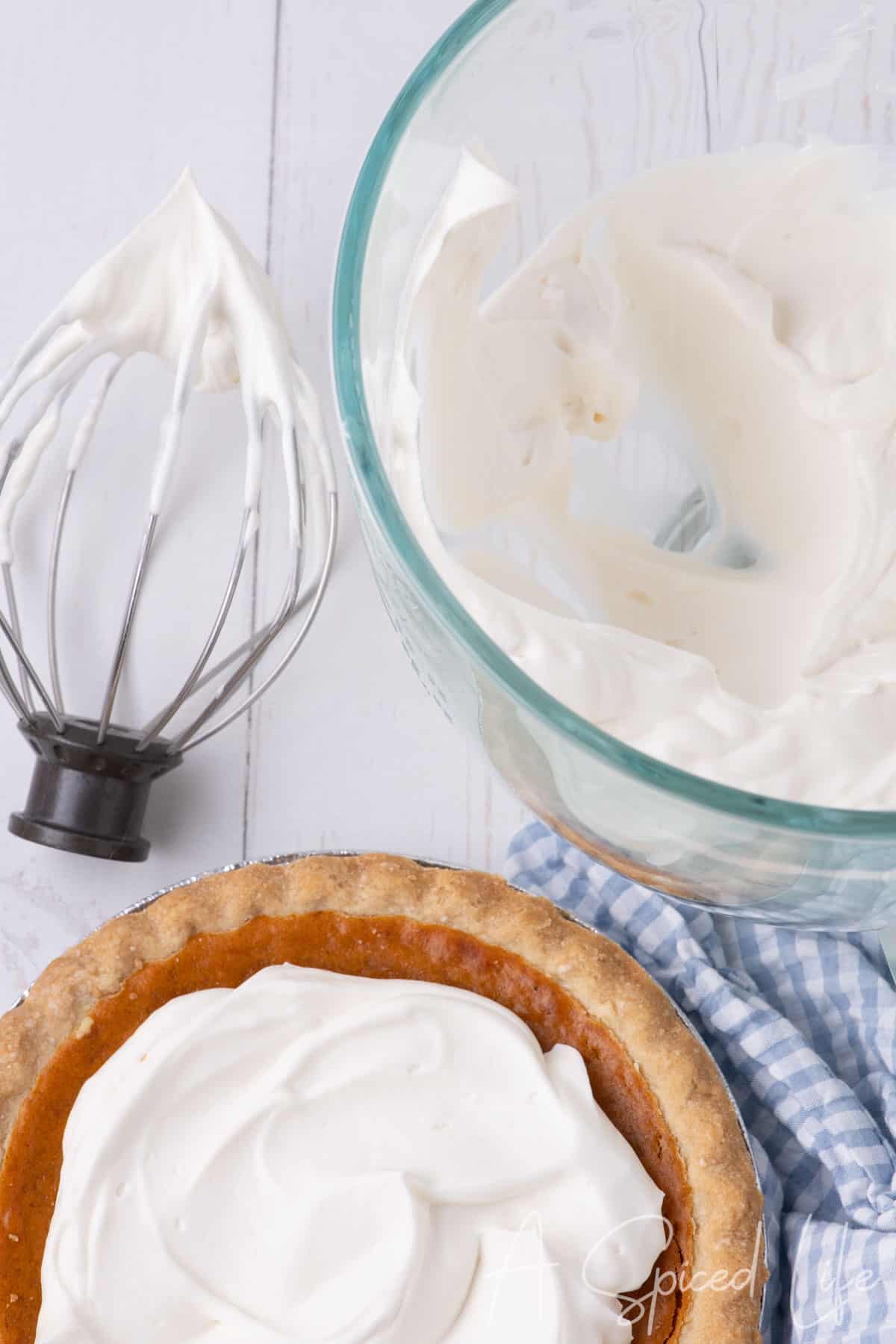 Glass bowl of whipped Chantilly cream next to a whisk and a pumpkin pie topped with fluffy cream.