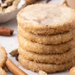 Close-up stack of spiced shortbread cookies showing crisp, sparkling sugar-coated edges with cinnamon sticks nearby.