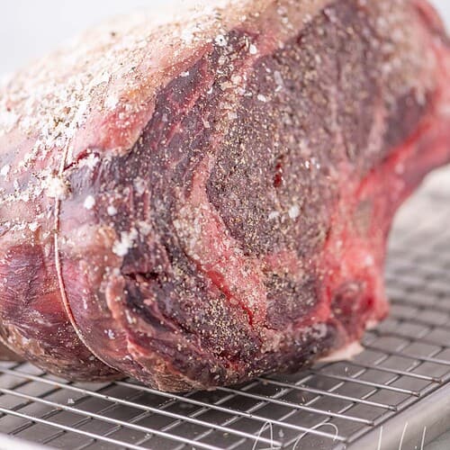 Hand sprinkling salt and pepper over a short dry aged prime rib roast on a wire rack.