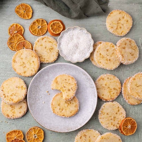 Overhead view of orange ginger cookies arranged with dried oranges and a plate on a neutral surface