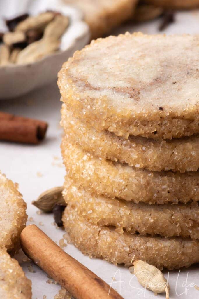 Close-up stack of spiced shortbread cookies with crisp, sparkling sugar-coated edges and cinnamon sticks in the foreground.