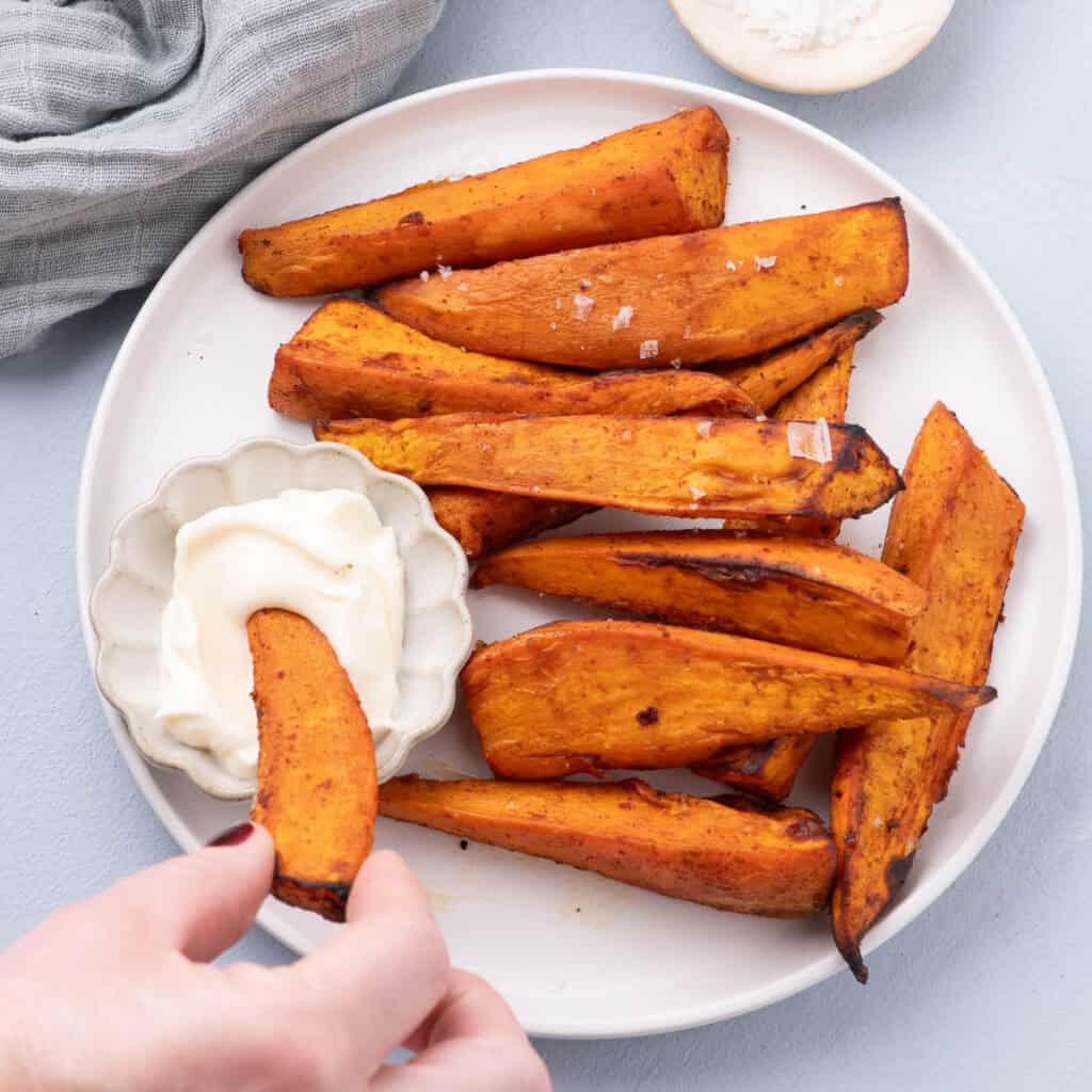 Crispy sweet potato wedges served on a plate with a creamy homemade mayo, one wedge being dipped.