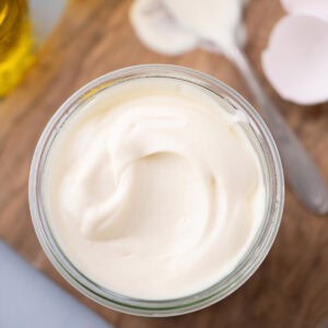 Top-down view of thick homemade mayonnaise in a small glass jar on a wooden board