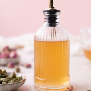 image of Rose simple syrup in glass dispenser surrounded by cardamom and rose petals