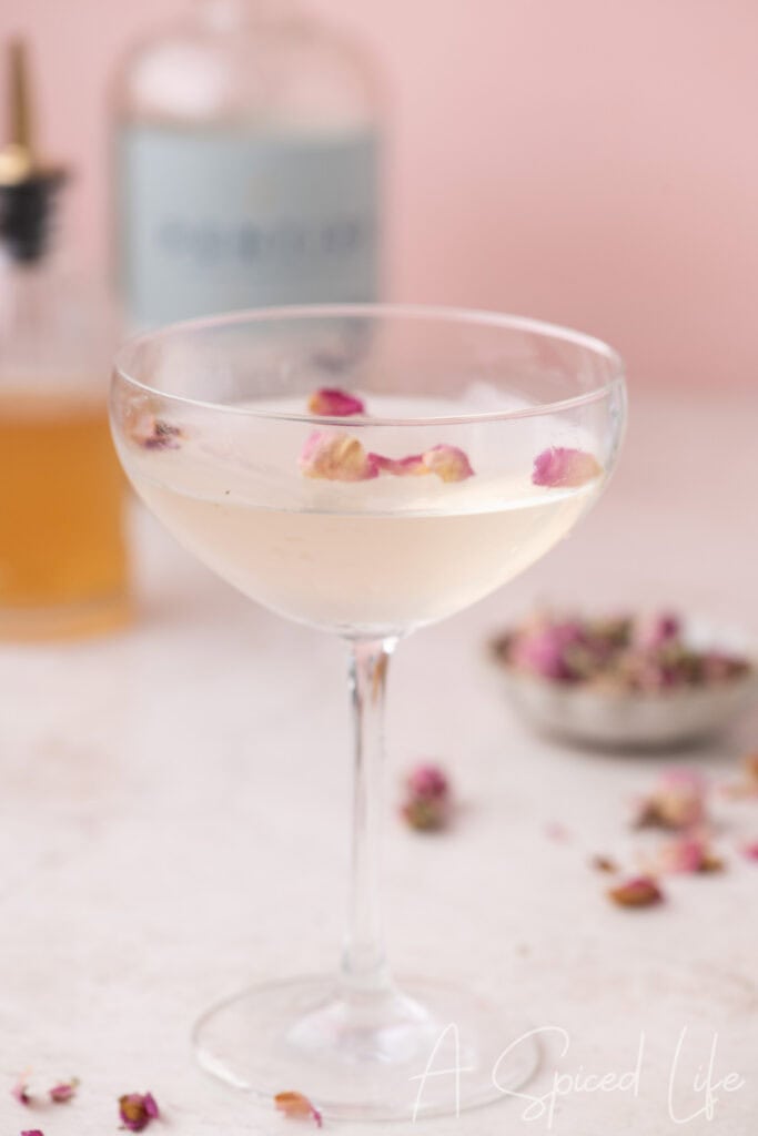Coupe glass filled with a clear rose mocktail topped with floating dried rose petals on a soft pink background