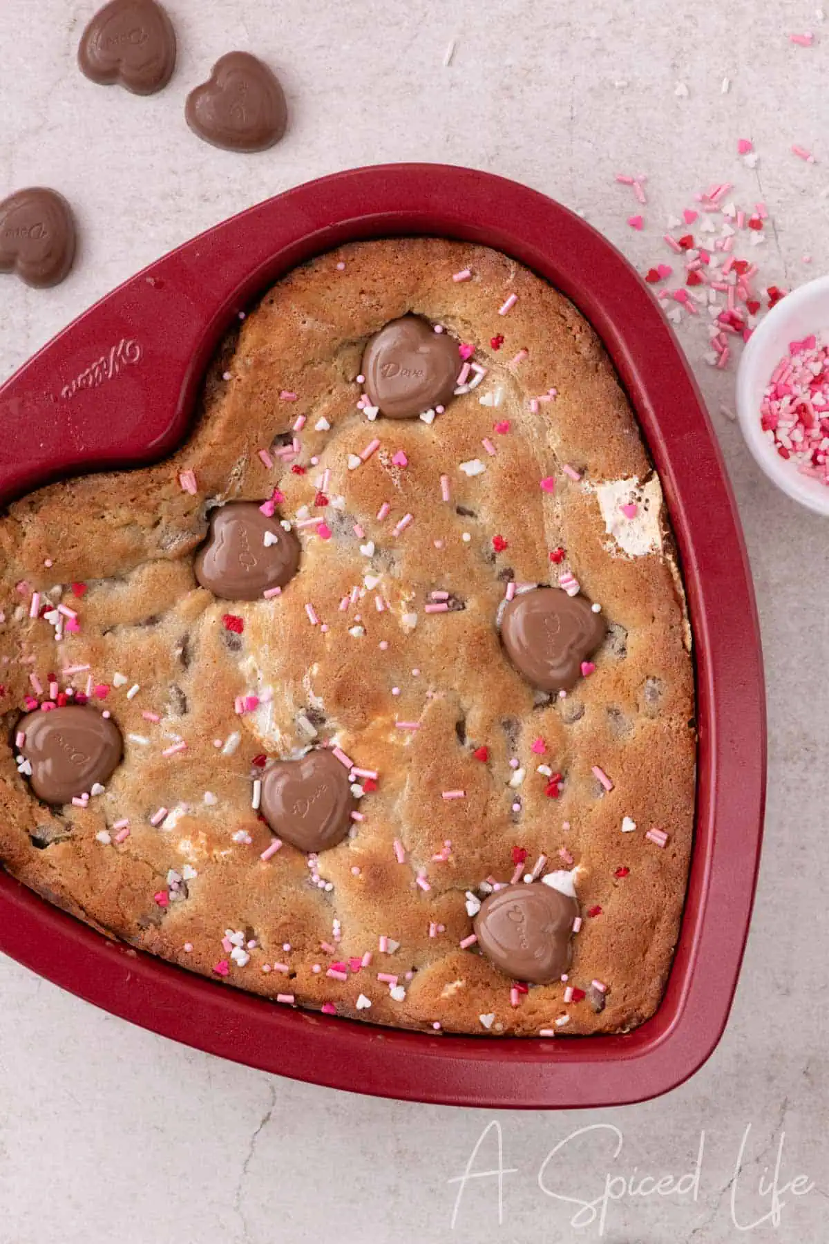 Golden heart-shaped cookie with melted chocolate hearts and pink sprinkles in a red baking pan