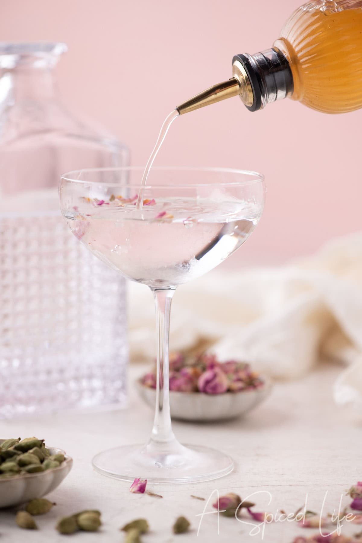 Syrup being poured from a bottle into a coupe glass with rose petals and crushed cardamom on a pink background