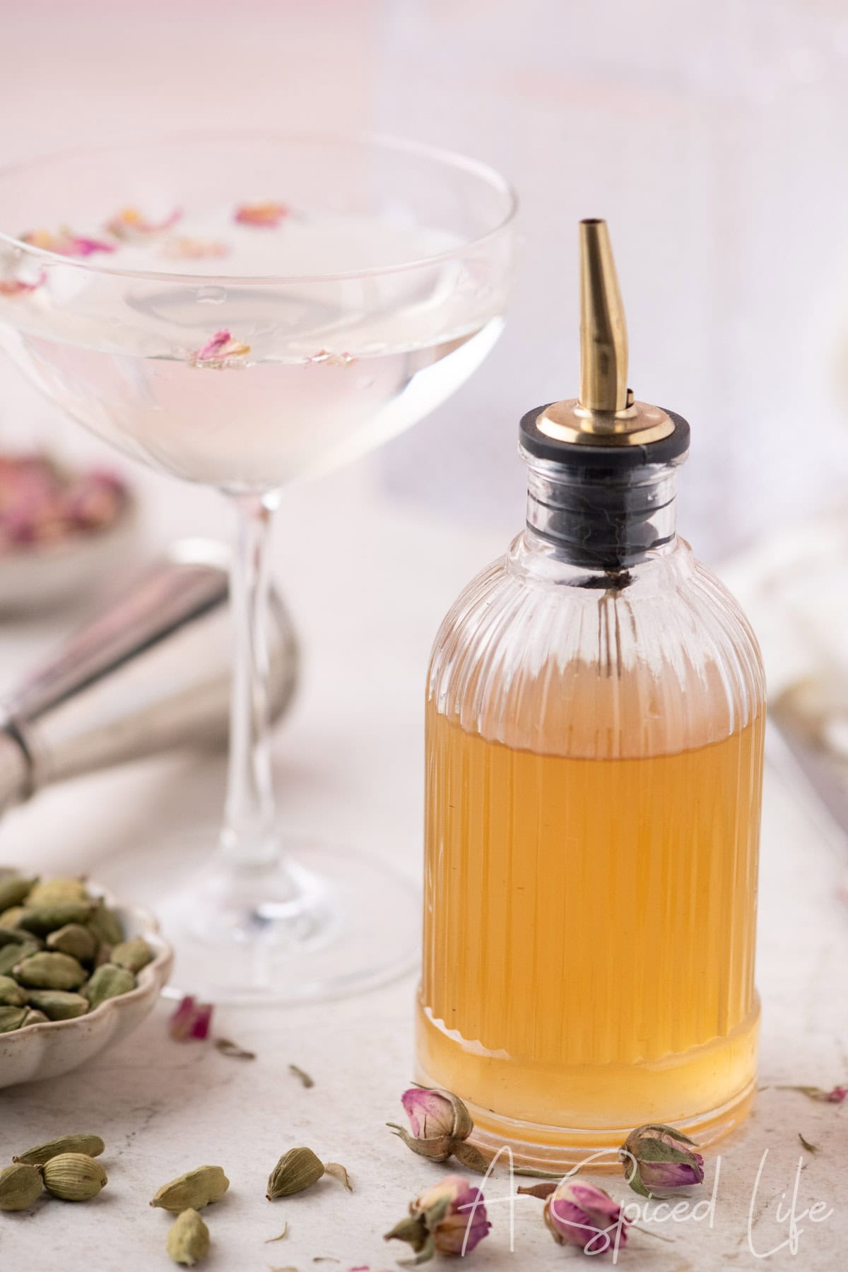 Bottle of golden floral syrup beside a coupe glass drink with cardamom pods and rose petals on a soft pink background
