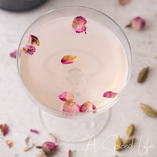 Overhead cocktail glass with floating rose petals and cardamom pods on a light surface