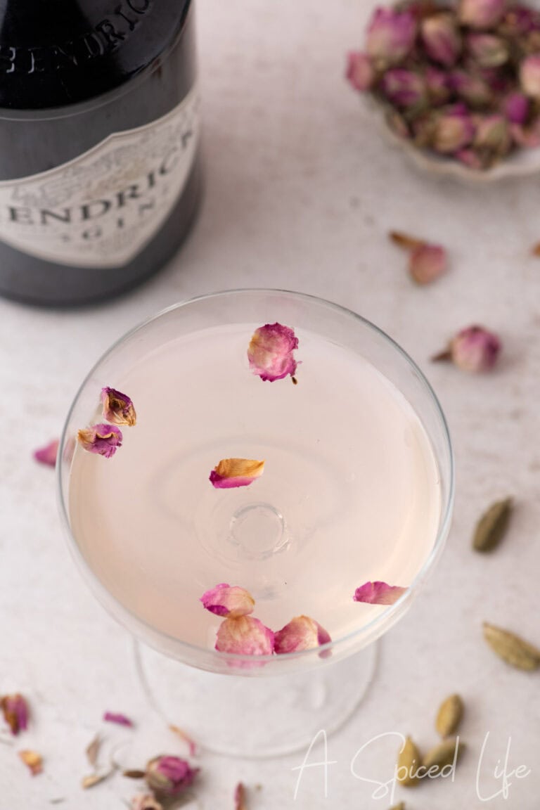 Overhead cocktail glass with floating rose petals and cardamom pods on a light surface
