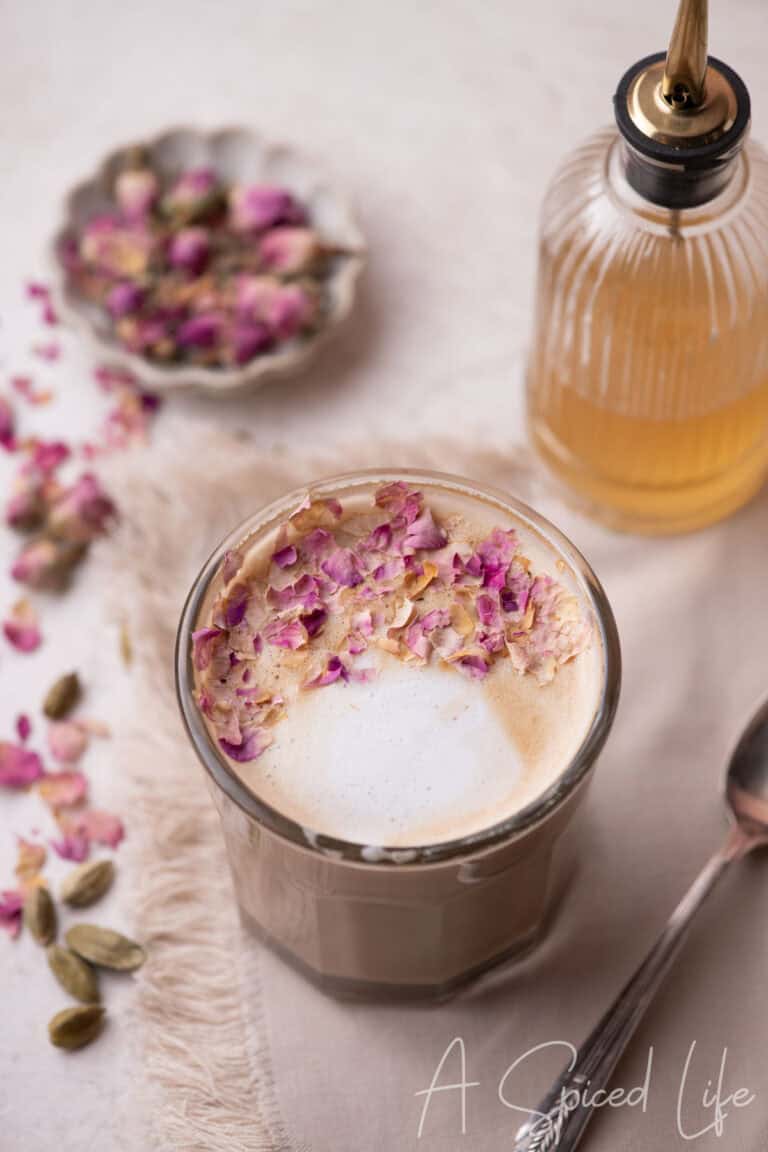 Glass of rose water latte topped with crushed rose petals, with rose cardamom syrup bottle in the background.