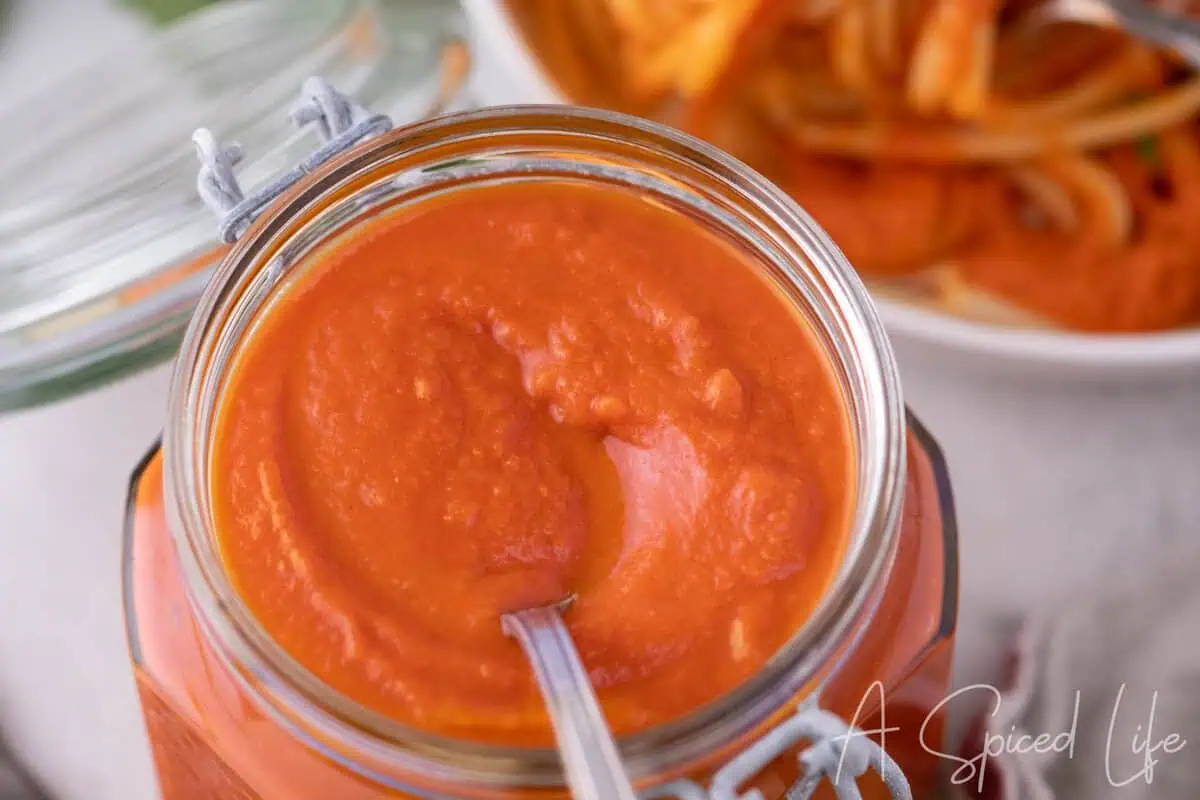 Jar of smooth San Marzano tomato sauce with a spoon and sliced tomatoes in the background.